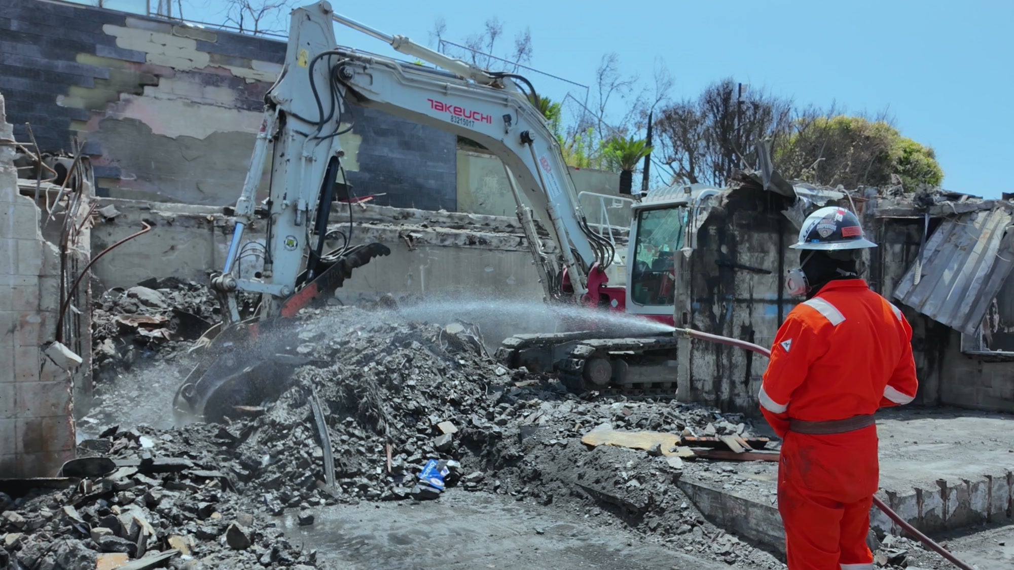 Excavator clearing fire-damaged masonry while a worker suppresses dust